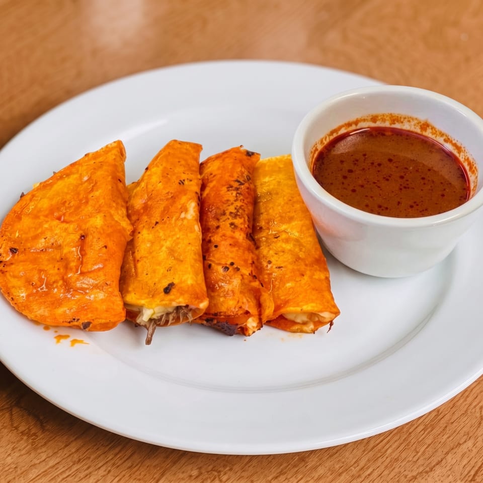Four crispy, orange-hued tacos with a side of red dipping sauce on a white plate.